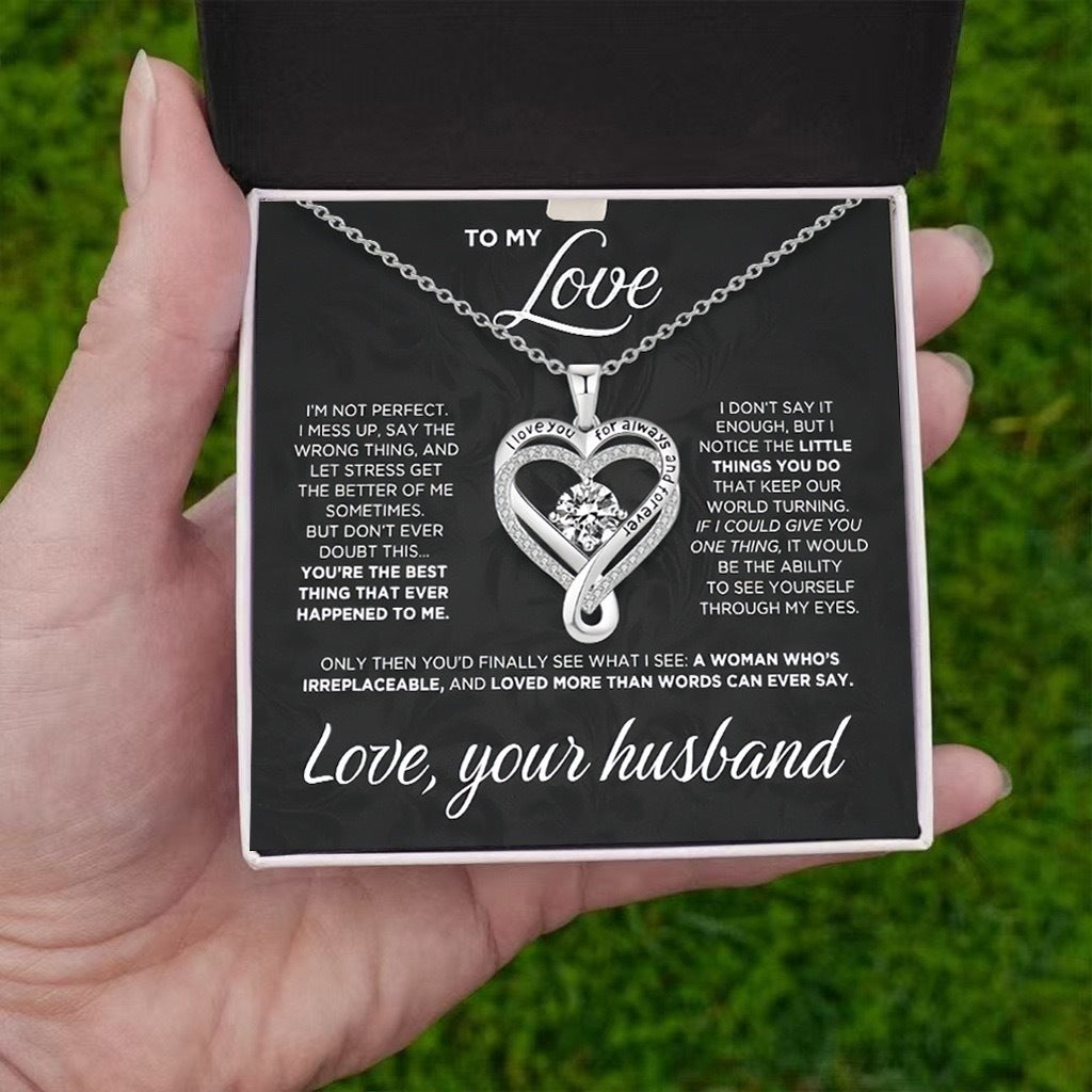 Heart-shaped necklace in a box with a sentimental message, held by a hand against a grassy background.